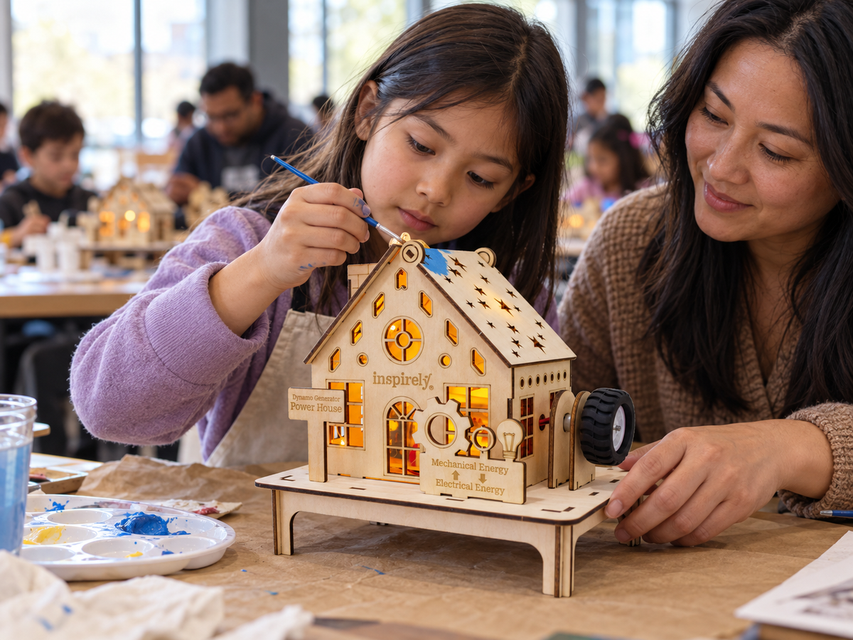 A woman and a young girl painting an Inspirely wooden STEM house model together at a craft table, with other people and tables visible in the blurred background.